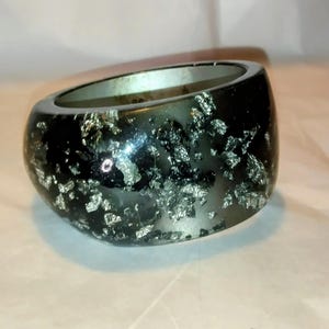 May include: A wide, thick bangle bracelet with a smooth, dark, semi-translucent resin base. The surface is embedded with numerous irregular, reflective silver-colored metallic flakes that create a high-contrast, shimmering, and textured appearance.