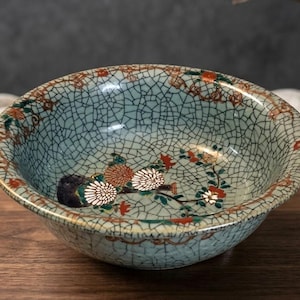 May include: A round ceramic bowl featuring a light blue crackle glaze finish. The interior and rim are decorated with hand-painted floral motifs in shades of orange, green, and white, accented with gold-coloured details throughout the design.