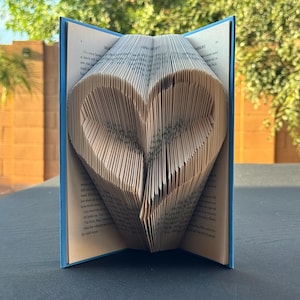 Peut inclure: Un livre dont les pages sont pliées pour former un cœur en trois dimensions au centre. Le livre possède une couverture bleue et les pages intérieures présentent du texte noir imprimé. Le pliage crée un motif de cœur symétrique visible.