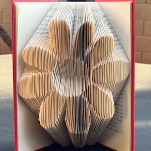 May include: A book with pages folded to create a three-dimensional flower shape. The book has a red cover and the interior pages are cream-coloured with black printed text. The folded pages form a central circular core with eight distinct petals.