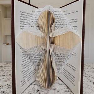 Op de afbeelding: Een boeksculptuur gemaakt van een hardcover boek met donkerbruine kaften. De pagina's zijn nauwkeurig gevouwen om een driedimensionale kruisvorm in het midden te vormen, waarbij de witte papierranden een gelaagd, geometrisch patroon creëren.