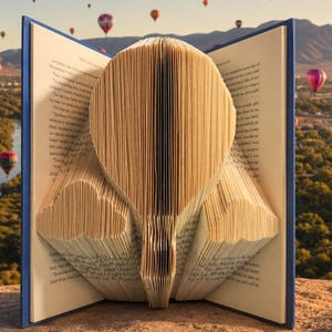 Puede incluir: Un libro con páginas dobladas para crear una escultura tridimensional con forma de globo aerostático. El libro tiene una cubierta azul y las páginas interiores son de color crema con texto negro impreso, mostrando la técnica de plegado.