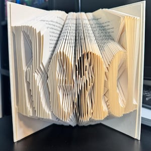 Può includere: Una scultura di libro con pagine piegate per formare la parola READ. Le pagine di carta color crema sono piegate con precisione per creare lettere tridimensionali che risaltano sulle copertine rigide color crema del libro aperto.