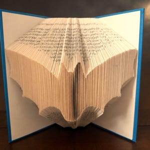 Puede incluir: Una escultura de libro con páginas dobladas para crear la silueta distintiva de un murciélago. El libro tiene una cubierta azul brillante y páginas blancas, con los bordes doblados formando las alas y el cuerpo del murciélago en el centro.