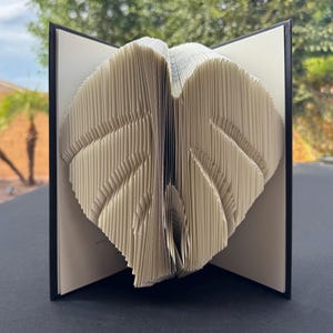 Puede incluir: Una escultura de libro con páginas dobladas para crear la forma de una hoja de monstera grande. El libro tiene una cubierta oscura y páginas de color crema, con pliegues intrincados que forman el patrón característico de la hoja en vertical.