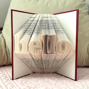 Puede incluir: Una escultura de libro con páginas dobladas que forman la palabra hello en minúsculas. El libro presenta una cubierta de color rojo intenso que enmarca las páginas de papel blanco, las cuales están plegadas con precisión para crear el texto 3D.