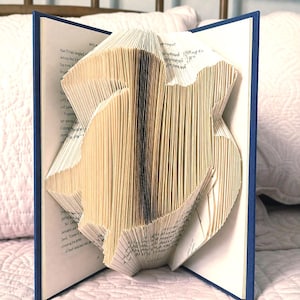 May include: A book with folded pages creating a three-dimensional silhouette of a crest. The hardback book features a dark blue cover and white paper pages with black printed text. The pages are precisely creased to form a central emblem shape.