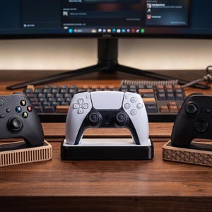 Puede incluir: Dos mandos de juego inalámbricos negros sobre soportes de madera clara con listones verticales y patrones geométricos, junto a un mando inalámbrico blanco en una base de carga negra, situados sobre una superficie de escritorio de madera oscura.