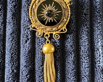 May include: A gold-tone brooch featuring a circular black center with a sunburst design and a central black stone. The frame has ornate scrollwork, small red and black accents, and a dangling gold-tone metal tassel suspended from the bottom.