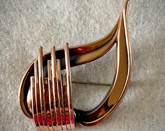 Puede incluir: Un broche de metal en tono cobre con un marco curvo en forma de lágrima. El lado izquierdo del broche está adornado con una serie de cinco barras verticales paralelas de diferentes alturas que se superponen al marco principal, creando un diseño lineal.