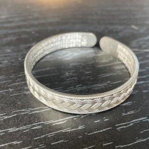 May include: A silver-toned open cuff bracelet featuring a detailed braided pattern along the outer band. The interior surface of the metal cuff is engraved with small, repeating text characters throughout the entire length of the inner band.