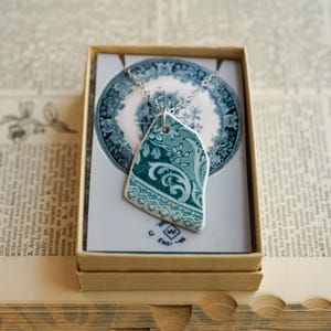 May include: A pendant necklace featuring a shard of teal and white floral patterned ceramic porcelain. The pendant hangs from a silver-toned chain and is displayed on a card printed with a matching circular plate design and the text SHAW ENGLAND.