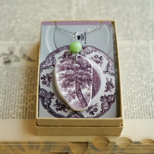 May include: A pendant necklace featuring a shard of white ceramic with a purple transferware tree design, accented by a single round light green bead. The pendant hangs from a silver-toned chain and is displayed on a card inside a cardboard box.