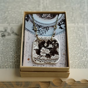 May include: A square ceramic pendant featuring a dark brown floral transferware pattern on a white background, suspended from a bronze-toned metal chain with small clear beads, presented inside a light brown cardboard gift box.