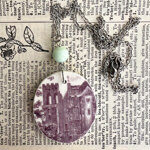 May include: A circular pendant necklace featuring a white ceramic shard with a purple transferware print of a historic building. The pendant is attached to a silver-toned metal chain by a single pale green bead and a small metal jump ring.