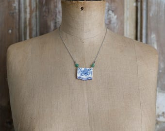 May include: A necklace featuring a rectangular ceramic pendant with a blue and white floral pattern. The pendant is attached to a thin silver-toned metal chain, accented by two small green beads positioned on either side of the ceramic piece.