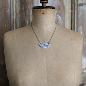 May include: A necklace featuring a semi-circular pendant made from white ceramic with a blue floral pattern. The pendant is suspended from a thin, dark metallic chain that rests against the neck of a beige fabric dress form mannequin.