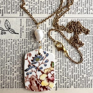 May include: A gold-toned chain necklace featuring a white, rectangular ceramic pendant with a floral pattern in blue, yellow, and brown. A single white, irregular-shaped pearl is positioned between the chain and the pendant.