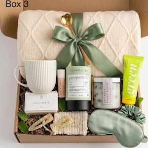 Op de afbeelding: Een geschenkdoos met het label Box 3 bevat een crèmekleurige gebreide deken met een saliegroen lint en een gouden lepel, een witte geribbelde mok, Katili Sabun zeep, grapefruit lavendel lippenbalsem, Tea With Tae thee, lavendel suikerscrub, een tube groene thee, twee haarclips, een textuurdoek en een saliegroen slaapmasker.
