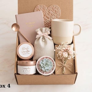 Op de afbeelding: Een geschenkdoos met een oudroze notitieboekje met de tekst Aesthetic Notes, een roségouden lepel, een beige keramische mok, een klein linnen zakje met trekkoord, twee ronde kaarsblikken Blush Vanilla en Lavender & Oud, een vetplant en gedroogde bloemen.