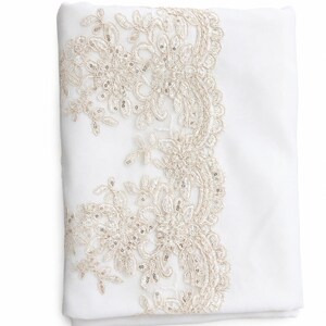 May include: A folded white fabric textile featuring an intricate beige lace border along one side. The lace pattern consists of floral motifs embellished with small, reflective silver sequins that catch the light, creating a textured appearance.