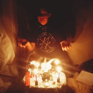 Puede incluir: Una camiseta negra con un diseño de sello circular blanco, usada por una persona detrás de una mesa pequeña. La mesa sostiene un cráneo de animal blanco, un pequeño caldero negro, varias velas encendidas e incienso, todo brillando en la luz.