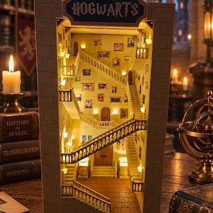 Può includere: Un diorama rettangolare da libreria che raffigura l'interno in miniatura di una scala di un castello in pietra con più livelli, ringhiere e quadri incorniciati alle pareti. La struttura è illuminata da luci calde e presenta un'insegna con la scritta HOGWARTS.