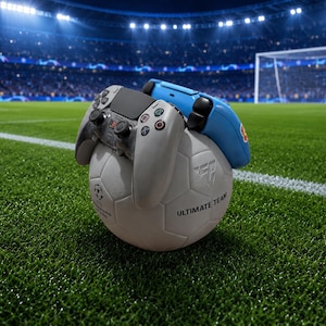 May include: A white football with the text ULTIMATE TEAM printed on its side, featuring a light grey game controller and a blue game controller resting on top of it, set against a backdrop of a professional football stadium pitch.