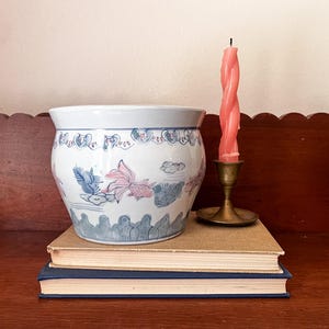 Puede incluir: Una maceta de cerámica blanca con cuerpo redondeado que presenta motivos de peces y nubes pintados en azul y rosa. El borde y la base tienen patrones festoneados decorativos. Junto a la maceta hay una vela rosa retorcida en un candelabro.