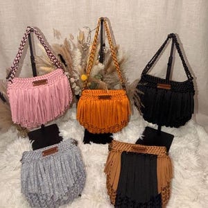 May include: Five crocheted handbags with fringe detailing. The collection includes solid pink, orange, and black bags, a heather gray bag, and one bag featuring a color-blocked design of tan and black. Each bag has a braided handle and a small label.