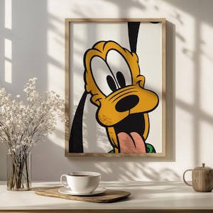 Op de afbeelding: Een ingelijste muurkunstprint met een close-up illustratie van het stripfiguur Pluto. De hond heeft een gele vacht, grote witte ogen met zwarte pupillen, een zwarte neus en een open mond met een roze tong die uitsteekt tegen een witte achtergrond.