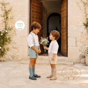 May include: Two children wearing white long-sleeved shirts with suspender shorts, one in light blue and one in beige, paired with knee-high socks and loafers. The child on the right holds a small bouquet of white flowers. Text reads SIZES 1-8 y.