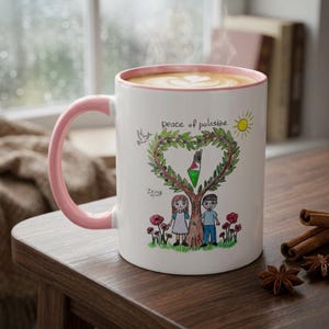 May include: A white ceramic mug with a light pink handle and rim, filled with coffee. The mug features a colorful hand-drawn illustration of a tree shaped like a heart, a dove, a sun, and two children, with the text "peace of palestine" and "Zeina".