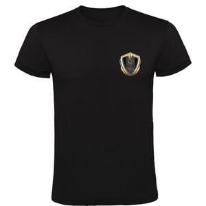 Puede incluir: Una camiseta negra de manga corta con un emblema en forma de escudo dorado y negro en el pecho izquierdo. El emblema muestra un gráfico de cobra estilizado y el texto SHELBY en letras negras, centrado dentro del diseño de borde dorado.