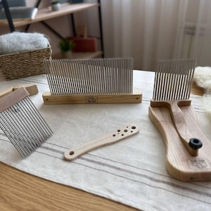 May include: A set of wooden wool combing tools including a long rectangular comb with metal tines, a smaller handheld comb with a wooden handle, a separate wooden handle with circular holes, and a wooden tool with a black knob and metal tines.