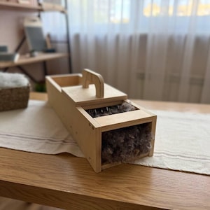Puede incluir: Una cardadora de lana rectangular de madera clara, fabricada en contrachapado, con un asa superior y una tapa deslizante. El dispositivo contiene una masa de fibras de lana cruda de color gris parduzco visible en el mecanismo de dientes metálicos.