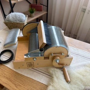 Puede incluir: Una cardadora de tambor de madera para procesar fibras con manivela, dientes metálicos en tambores giratorios y un cepillo de madera. El dispositivo tiene un marco de madera clara con un pequeño logotipo y una herramienta de cardado manual.