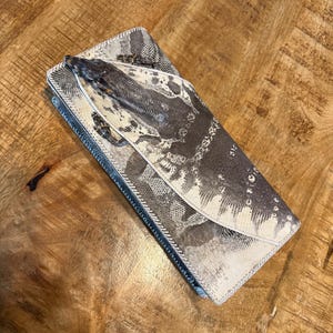 May include: A rectangular wallet featuring a textured, grey and white snakeskin-patterned exterior. The wallet has a flap closure with a distinct, raised, three-dimensional lizard figure attached to the front surface, finished with white piping.