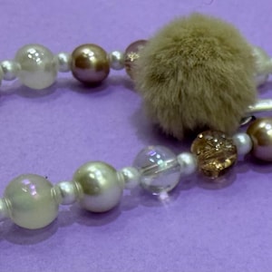 Puede incluir: Una correa de cuentas para la muñeca con una mezcla de cuentas blancas, iridiscentes y de color bronce metálico, acentuada con un suave pompón de piel sintética beige y un dije de concha plateado, con un cierre de mosquetón de metal.