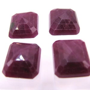 May include: Four rectangular faceted gemstones in a deep magenta-red color. Each stone features a step-cut design with a raised central table and beveled edges, displaying a translucent quality and a polished surface finish across all four pieces.