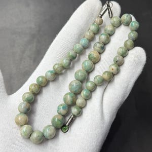 May include: A beaded necklace featuring round, polished stones in variegated shades of pale green, cream, and soft blue. The beads are strung together and secured with a simple silver-toned metal clasp at the ends of the strand.
