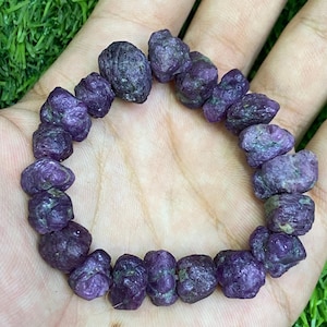 Puede incluir: Una pulsera compuesta por numerosas cuentas de piedras preciosas en bruto, sin pulir, de color púrpura intenso y magenta oscuro. Cada cuenta presenta una superficie irregular, texturizada y rugosa, ensartadas en círculo.