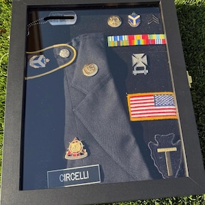 May include: A black shadow box display case containing a dark blue military uniform jacket, a name plate reading CIRCELLI, a US flag patch, various metal pins, ribbons, a dog tag, and an arrowhead-shaped patch with a T, all arranged on a black background.
