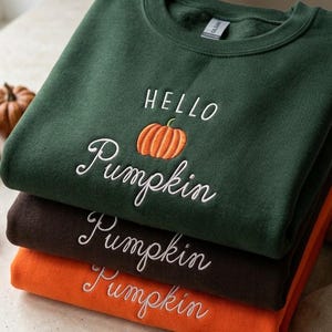 May include: A stack of three crewneck sweatshirts in dark green, brown, and orange. The top green sweatshirt features the embroidered text "HELLO" above an orange pumpkin icon and the word "Pumpkin" below. The other two sweatshirts feature "Pumpkin" embroidered in white script.