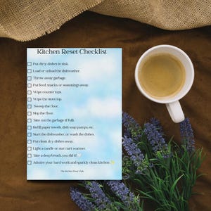 May include: A rectangular Kitchen Reset Checklist printed on a light blue sky-patterned background. The list includes tasks like putting dishes in the sink and wiping counters. A white ceramic mug of coffee and purple lavender sprigs are nearby.