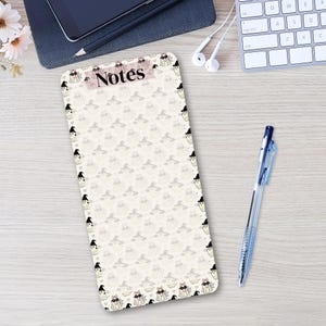 May include: A rectangular notepad with rounded corners featuring a repeating pattern of small ghosts wearing witch hats and glasses. The word Notes is printed in black at the top center against a light pink rectangular background.