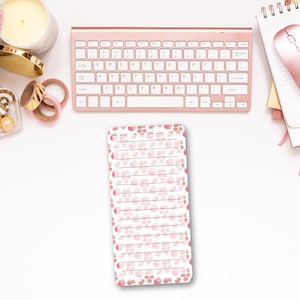 May include: A rectangular notepad featuring a repeating pattern of small pink cherries with green stems and leaves. The notepad has horizontal lines for writing and is positioned below a rose gold wireless keyboard, a candle, tape rolls, and a notebook.