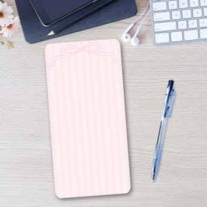 Può includere: Un blocco note rettangolare caratterizzato da un motivo a righe verticali rosa chiaro e bianco con una delicata illustrazione a linee di un fiocco al centro in alto. La carta presenta angoli arrotondati e una finitura liscia.