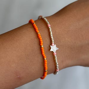 Può includere: Due braccialetti di perline indossati insieme. Un braccialetto presenta piccole perline arancioni vivaci intervallate da minuscoli distanziatori metallici dorati. Il secondo braccialetto è composto da perline metalliche argentate, accenti rosa chiaro e un ciondolo a forma di stella bianca.