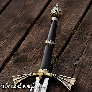 Peut inclure: Une épée avec une poignée noire, un pommeau couleur laiton orné d'un motif triangulaire et une garde aux extrémités évasées et texturées. La lame présente un motif en acier Damas et le texte The Lord Knight est visible.